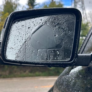 Pode incluir: Um espelho retrovisor de carro preto com gotas de água na superfície. O espelho reflete um céu nublado e árvores.