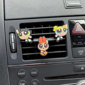 Op de afbeelding: Drie luchtverfrissers voor in de auto met de tekenfilmfiguren Blossom, Bubbles en Buttercup van The Powerpuff Girls, bevestigd aan een ventilatierooster. De personages zijn in hun kenmerkende kleuren roze, blauw en groen. Het dashboard van de auto is zichtbaar op de achtergrond.