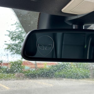 Puede incluir: Un espejo retrovisor en un coche con una pequeña pegatina de extraterrestre en el cristal.