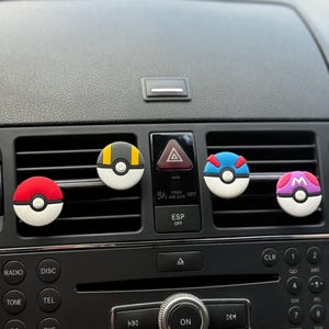 May include: A car dashboard featuring five air vent clip-on accessories. The accessories are round and feature various colours and designs, including a red and white ball, and a yellow and black ball.