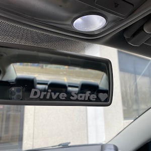 Può includere: Uno specchietto retrovisore con un adesivo grigio "Drive Safe" con un simbolo di cuore all'interno di un'auto.