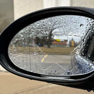 May include: A close-up of a car's side view mirror with a star-shaped rain guard. The mirror is covered in water droplets.