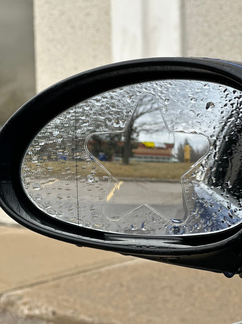 May include: A close-up of a car's side view mirror with a star-shaped rain guard. The mirror is covered in water droplets.