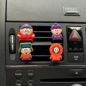May include: Four colourful cartoon character air vent charms. The characters are wearing red, blue, green, and orange outfits. The charms are attached to a car's air vent.