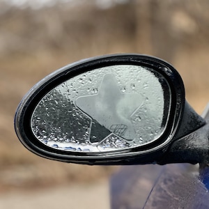 May include: A black car side mirror with a clear star-shaped sticker on the glass. The mirror is covered in water droplets.