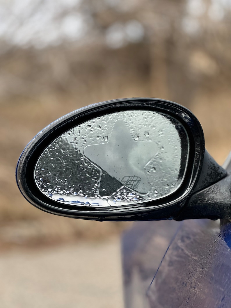 May include: A black car side mirror with a clear star-shaped sticker on the glass. The mirror is covered in water droplets.