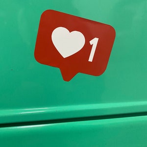 May include: A red sticker in the shape of a speech bubble, featuring a white heart and the number 1, set against a teal background. The design is reminiscent of a social media 'like' icon.