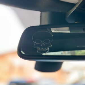 Könnte beinhalten: Schwarzer Rückspiegel mit silbernem Totenkopf-Emblem. Der Totenkopf hat detaillierte Augenhöhlen, eine Nasenhöhle und eine Zahnreihe. Der Spiegel ist in einem Fahrzeug montiert.