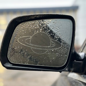 May include: A black car side mirror with water droplets on the surface. A white sticker of a planet with rings is visible on the mirror.
