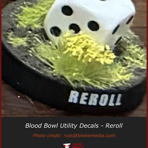 May include: A white six-sided die with black pips resting on a circular black base covered in textured green grass-like material. The front edge of the black base features the word REROLL printed in white capital letters in a bold font.
