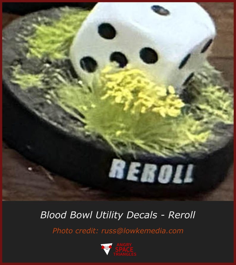May include: A white six-sided die with black pips resting on a circular black base covered in textured green grass-like material. The front edge of the black base features the word REROLL printed in white capital letters in a bold font.
