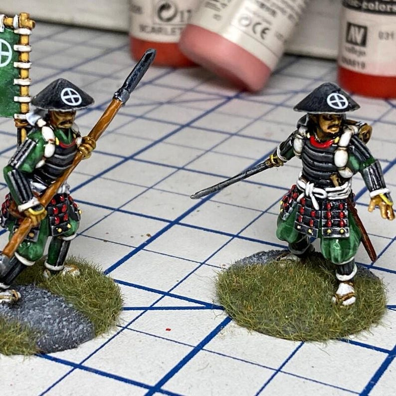 Op de afbeelding: Twee miniatuur samoerai-figuurtjes op ronde, met gras bedekte voetstukken. De figuren dragen zwart-groene harnassen, witte sjerpen en zwarte kegelvormige hoeden met witte cirkelvormige symbolen. E&eacute;n houdt een lange houten speer, de andere een zwaard.