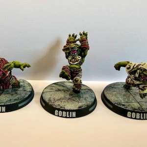 May include: Three painted miniature goblin figurines standing on circular bases with the word GOBLIN printed on the front. The figures feature green skin, dynamic poses, and detailed armour in red, white, and metallic tones, set on stone-textured bases.