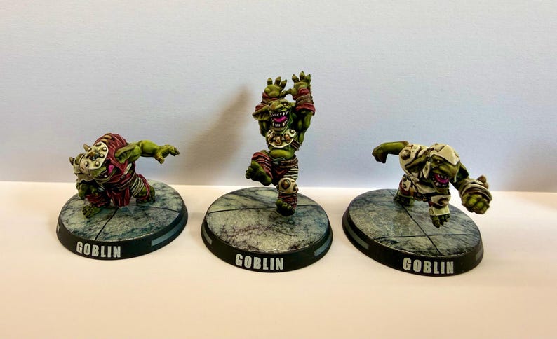 May include: Three painted miniature goblin figurines standing on circular bases with the word GOBLIN printed on the front. The figures feature green skin, dynamic poses, and detailed armour in red, white, and metallic tones, set on stone-textured bases.