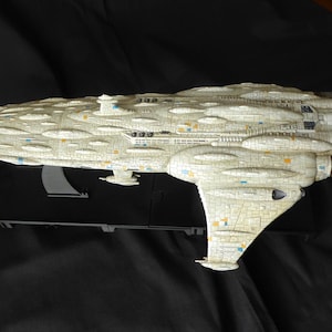 May include: A detailed scale model of a Mon Calamari star cruiser featuring an elongated, off-white hull with a textured, organic surface pattern. The spacecraft includes multiple rear engine thrusters and small blue and orange accent markings.