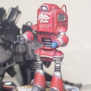 Op de afbeelding: Een miniatuur robotfiguur geschilderd in rood met zilveren accenten, voorzien van de witte tekst Nuka Cola op de borst. De robot heeft een hoekig hoofd, een veerachtige romp en twee armen waarvan &eacute;&eacute;n hand is uitgestrekt op een ronde voet.