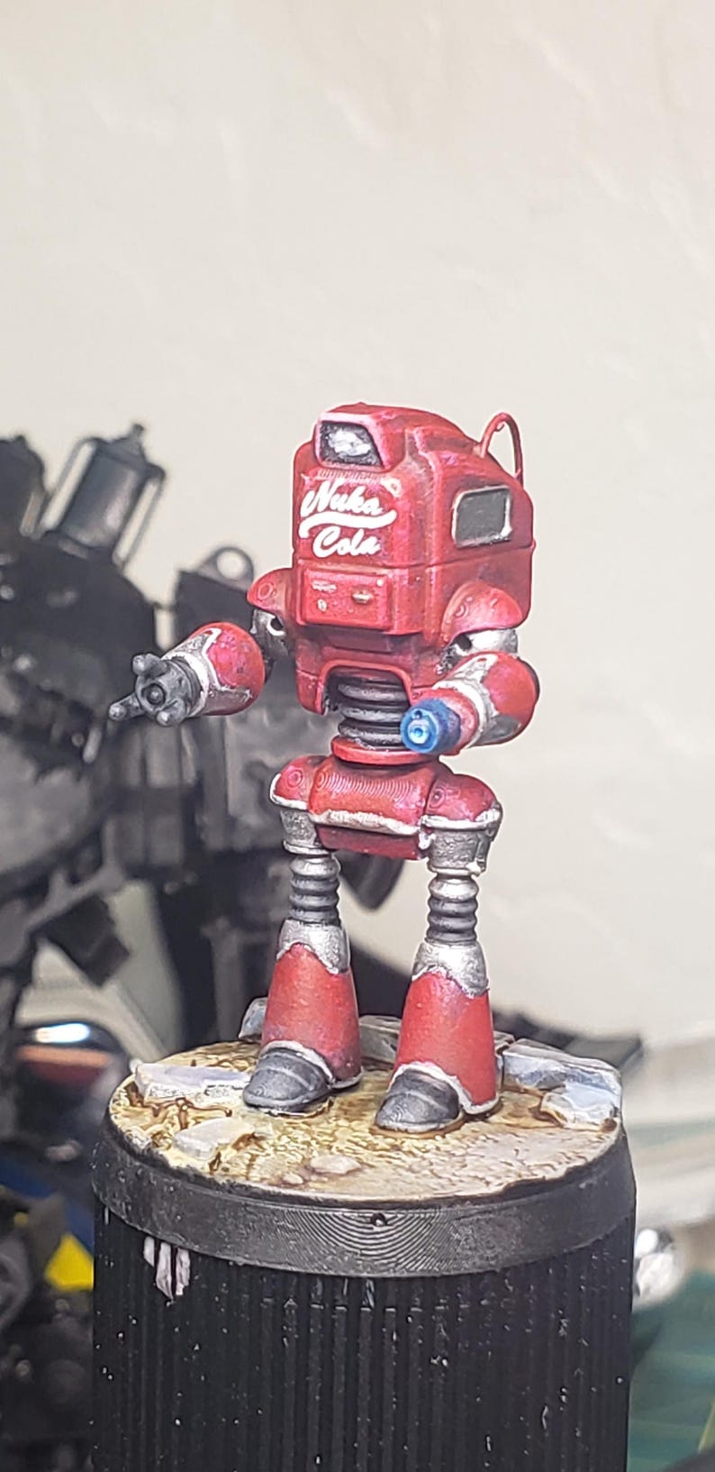 Op de afbeelding: Een miniatuur robotfiguur geschilderd in rood met zilveren accenten, voorzien van de witte tekst Nuka Cola op de borst. De robot heeft een hoekig hoofd, een veerachtige romp en twee armen waarvan &eacute;&eacute;n hand is uitgestrekt op een ronde voet.