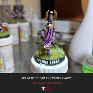 May include: A miniature Blood Bowl High Elf Phoenix Guard figure standing on a circular base with green grass texture. The figure wears a silver helmet with gold wings, a purple cape, and holds a football. The base is labelled PHOENIX GUARD.