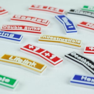 May include: A collection of colorful, rectangular game cards with various text and symbols. The cards feature words like "Flying", "Lifelink", "Indestructible", "Double Strike", "Loaded", "Ward 1", "Death touch", "Hexproof", and "+1/+1".