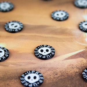 May include: Close-up of black and white round tokens with the text "Keep Up" and a skull graphic. The tokens are scattered on a brown surface.