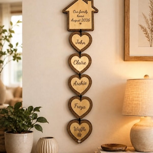 May include: A vertical wooden wall hanging featuring a house-shaped top piece engraved with "Our family home August 2026" followed by five connected heart-shaped wooden plaques engraved with the names John, Claire, Archie, Freya, and Waffle.