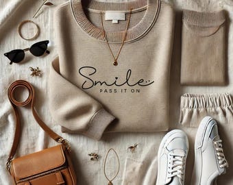 May include: A beige sweatshirt with the text "Smile... Pass it on" in black script. The sweatshirt is laid out on a white and beige textured surface with a brown leather purse, gold jewelry, and white sneakers.