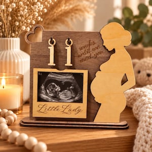 May include: A wooden pregnancy countdown sign featuring a silhouette of a pregnant person, two number 1s, and a frame holding an ultrasound image. The text reads "weeks until we meet you" and "Little Lady" in cursive script on a light wood frame.