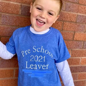May include: A young child wearing a blue t-shirt with the text "Pre School 2021 Leaver" in white. The child is smiling and standing in front of a red brick wall. The shirt has a white long-sleeved undershirt.