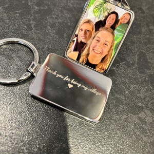 May include: Silver keychain with a photo insert featuring four women smiling. The back of the keychain is engraved with the text "Thank you for being my village" and a small heart.