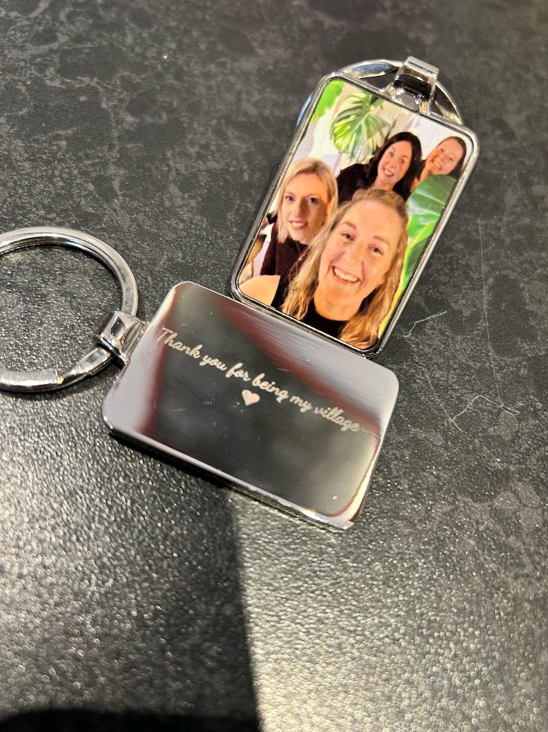 May include: Silver keychain with a photo insert featuring four women smiling. The back of the keychain is engraved with the text "Thank you for being my village" and a small heart.