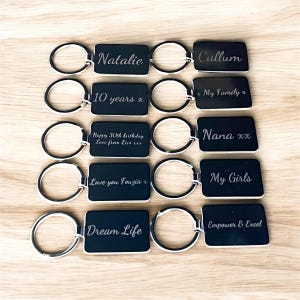 May include: Ten black metal keychains with silver rings. Each keychain has a rectangular tag with a different engraved message, including "Natalie", "Callum", "My Family x", "10 years x", "Happy 30th birthday Love from Love", "Nana xx", "Love you Fouzia x", "My Girls", "Dream Life", and "Empower & Excel".
