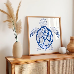 Puede incluir: Un cuadro con una ilustración de tortuga azul con patrones intrincados. La obra de arte se exhibe en un mueble de madera con frente tejido, junto a un jarrón con hierba de la pampa y otros elementos decorativos. La estética general es costera y bohemia.