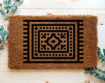 May include: A rectangular doormat made of natural coir fibers. The mat features a black geometric design in the center, resembling a rug, with a border of vertical lines. The design includes a central diamond shape and dotted details.