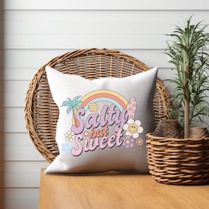 May include: A white throw pillow with a colorful design featuring a rainbow, palm trees, a surfboard, flowers, and the text "Salty but Sweet".