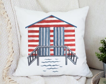 May include: A white cushion featuring a red and white striped beach hut illustration. The hut has blue doors and windows and a grey wooden deck with railings. The illustration is set against a blue water background.