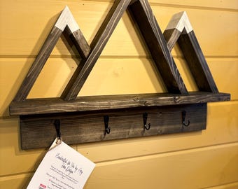 May include: Wooden mountain-shaped shelf with a white painted peak and four black hooks. The shelf is mounted on a yellow wall. A small white tag with the text "Handmade with Love by Amy, Shop Design" is attached to the shelf.