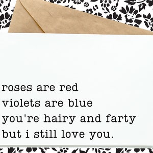 May include: A white greeting card with the text "roses are red, violets are blue, you're hairy and farty but I still love you." The card is inside a brown envelope, set against a black and white floral pattern.