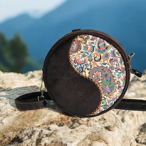 May include: A round brown and white patterned crossbody bag with a long strap. The bag has a yin yang design with one side being brown suede and the other side being a white fabric with a paisley pattern.