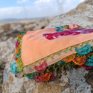 May include: A peach-colored fabric square with a floral print and a crocheted border. The border is made of green, blue, orange, and red yarn.