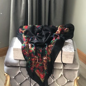 May include: A black patterned scarf with red, green, and gold accents is draped over a gray velvet ottoman with gold legs.