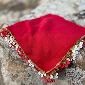 May include: A red square fabric with white and green crocheted trim. The fabric is folded and lying on a rock.