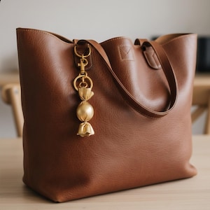 May include: A brown leather tote bag with a gold-colored candy charm hanging from the handle. The bag has a structured design with a top handle and a textured surface. The charm is a decorative accent.