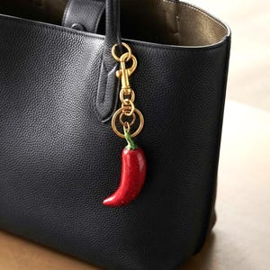 May include: A black textured leather tote bag with a gold-toned metal lobster clasp and ring keychain attached to the handle. The keychain features a glossy, bright red chili pepper charm with a small green stem at the top of the pepper.