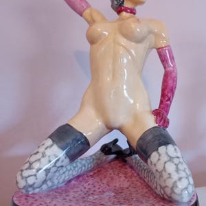 Könnte beinhalten: Keramikfigur einer knienden Frau mit nacktem Oberkörper und schwarzem Haar. Sie trägt rosa Handschuhe, ein Halsband und Strümpfe. Die Figur steht auf einem rosa und schwarzen Sockel.