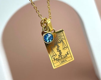 May include: A gold-toned necklace featuring a rectangular pendant engraved with a scale symbol and the word LIBRA. A small, round, faceted blue crystal charm is attached to the same jump ring as the pendant, hanging from a delicate gold chain.