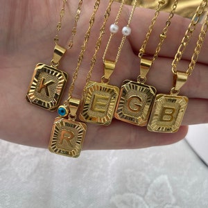 May include: Five gold necklaces with square pendants featuring the letters K, E, G, B, and R. The pendants have a textured, sunburst design. One necklace has a blue evil eye charm.