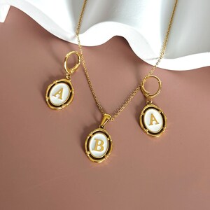 May include: Gold necklace and earrings set with white oval pendants featuring the letters A and B.