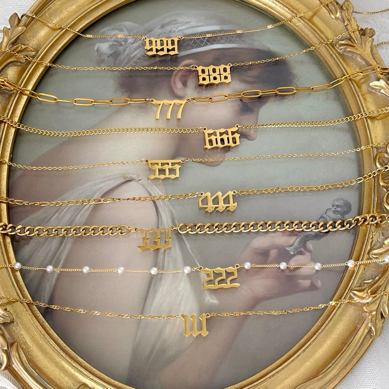 May include: A collection of nine gold-toned necklaces, each featuring a different number pendant in an old English font. The chains vary in style, including delicate links, paperclip links, curb chains, and a chain accented with small white pearls.