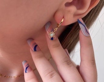 Pode incluir: Brincos de argola dourados com um charme de estrela pendurado que apresenta uma pedra de opala azul. Os brincos são mostrados contra um fundo de uma mão com esmalte de unha azul e rosa.