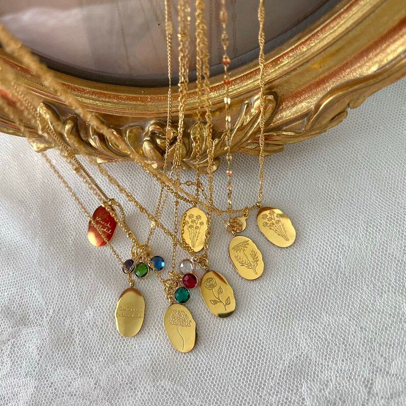 May include: A selection of gold necklaces with oval pendants. Some pendants feature floral engravings, while others have colourful gemstones or engraved text. The necklaces are displayed against a gold-framed background.